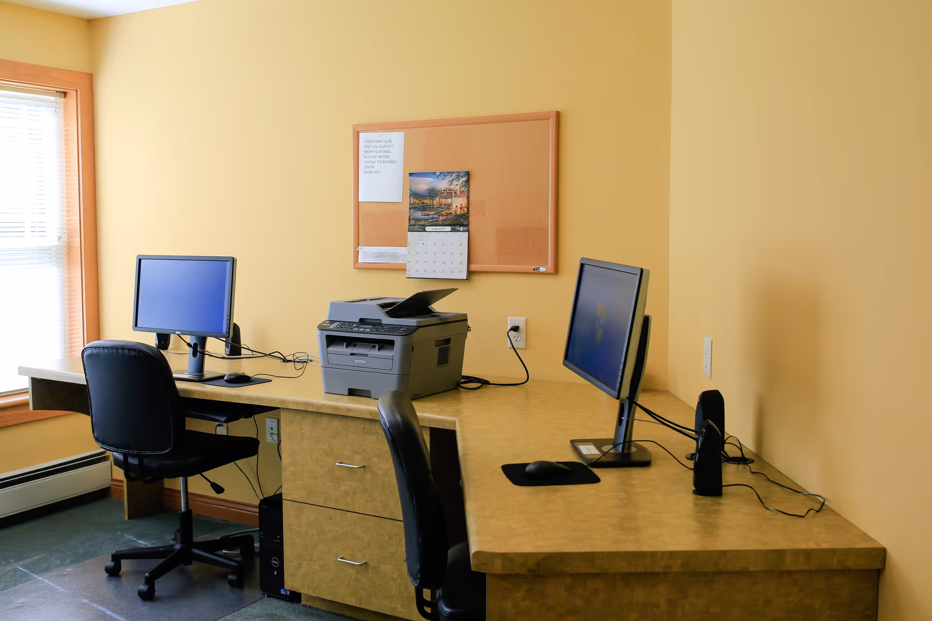 A small office area with two computer workstations, each with a monitor, keyboard, and mouse. There is a printer placed between the two workstations on a beige countertop. A cork bulletin board with a calendar and a notice is mounted on the yellow wall behind the desk. A window with blinds is on the left side, allowing natural light into the room.