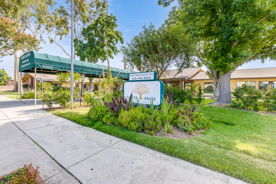 Exterior view of Santa Anita Assisted Living facility showing a green awning over the entrance with the facility name and address. There is a landscaped garden with various plants and trees surrounding a sign displaying the facility's name, address, and phone number. The building is single-story with large windows and a sidewalk leading to the entrance.