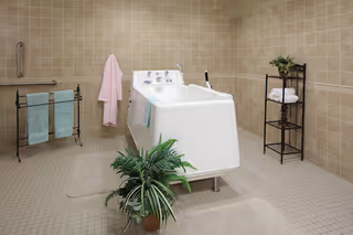 A spacious bathroom with beige tiled walls and floor featuring a white walk-in bathtub in the center. There are towels hanging on a black metal rack and a pink robe hanging on a wall hook. A black metal shelving unit holds neatly folded white towels and a small plant. A green potted plant is placed on the floor near the bathtub.