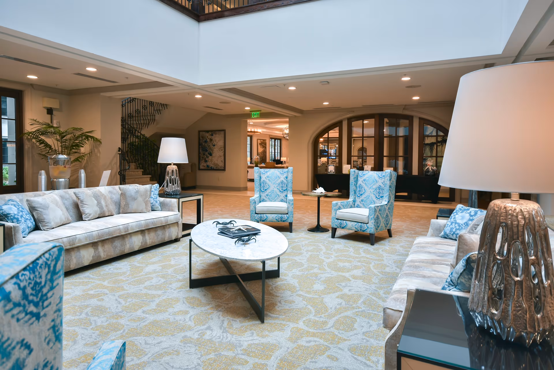 A spacious and well-lit living room area with two beige sofas adorned with blue and beige cushions, two blue patterned armchairs, a white oval coffee table with decorative items, and two modern table lamps. The room features a patterned carpet, a staircase with a wrought iron railing, and large windows and doors allowing natural light.