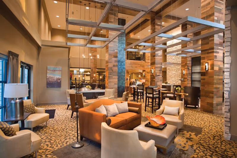 A spacious and well-lit common area in a nursing home featuring comfortable seating including a brown sofa and beige armchairs arranged around a coffee table with decorative items. The room has high ceilings with modern hanging light fixtures, wooden and stone accent walls, patterned carpet, and large windows with curtains allowing natural light.