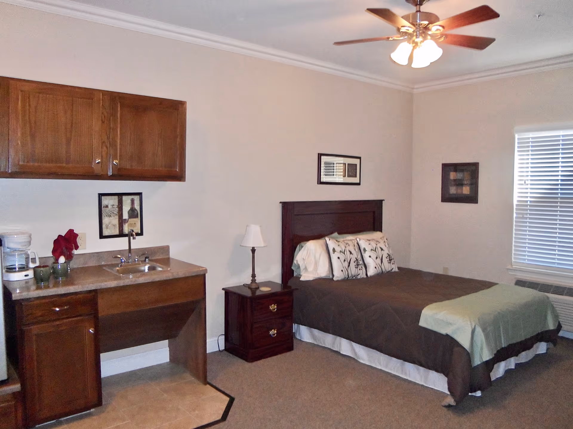 A cozy assisted living bedroom featuring a bed with decorative pillows and a green throw blanket, a wooden nightstand with a lamp, a window with blinds, and a small kitchenette area with a sink, countertop, coffee maker, and upper cabinets. The room has beige walls, carpeted flooring, and a ceiling fan with lights.