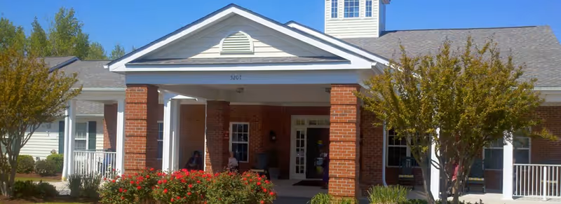 Front entrance of a brick senior living building with white columns, a covered portico, and landscaping under a clear blue sky.