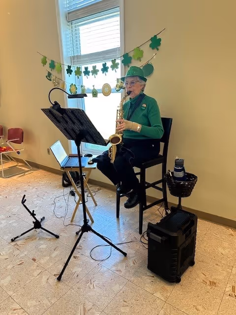 An elderly woman dressed in green, wearing a green hat with shamrock decorations, is sitting on a high chair playing a saxophone. She is positioned near a window decorated with a garland of green shamrocks. In front of her is a music stand with sheet music, and nearby are a microphone stand, a laptop, and a speaker on the floor. The room has beige walls and a tiled floor.