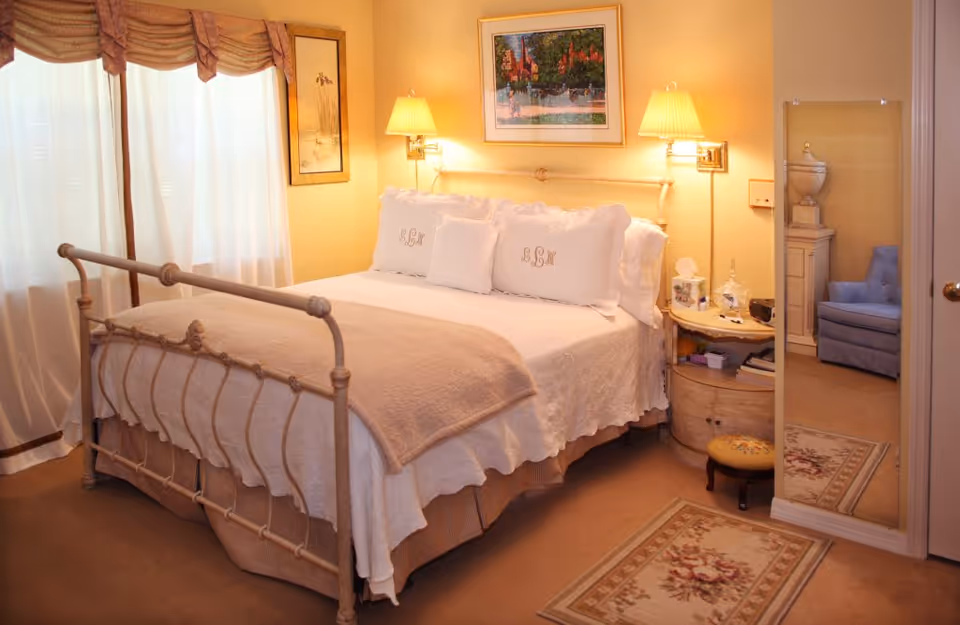 A cozy bedroom with a metal-framed bed covered in white bedding and beige blanket. The bed has three white pillows with monogrammed initials. On the right side, there is a round wooden nightstand with a lamp, tissue box, and other small items. A tall mirror leans against the wall next to the nightstand, reflecting a blue armchair and decorative urn. The room has beige walls, carpeted floor, floral rugs, and a window with sheer white curtains and a valance.