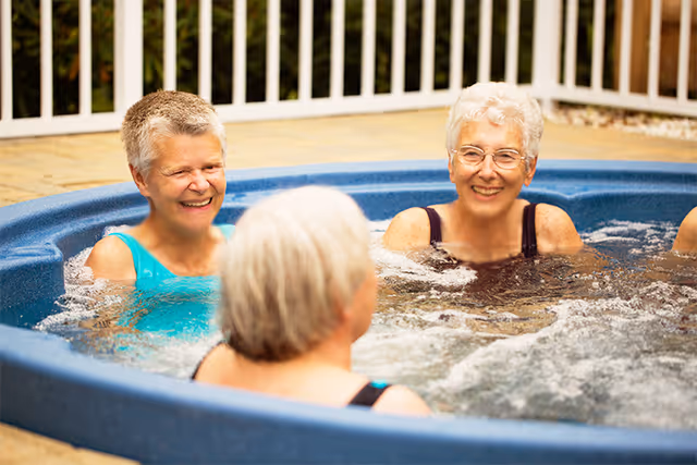 Three elderly women enjoying a conversation while sitting in a bubbling outdoor hot tub surrounded by a white fence.