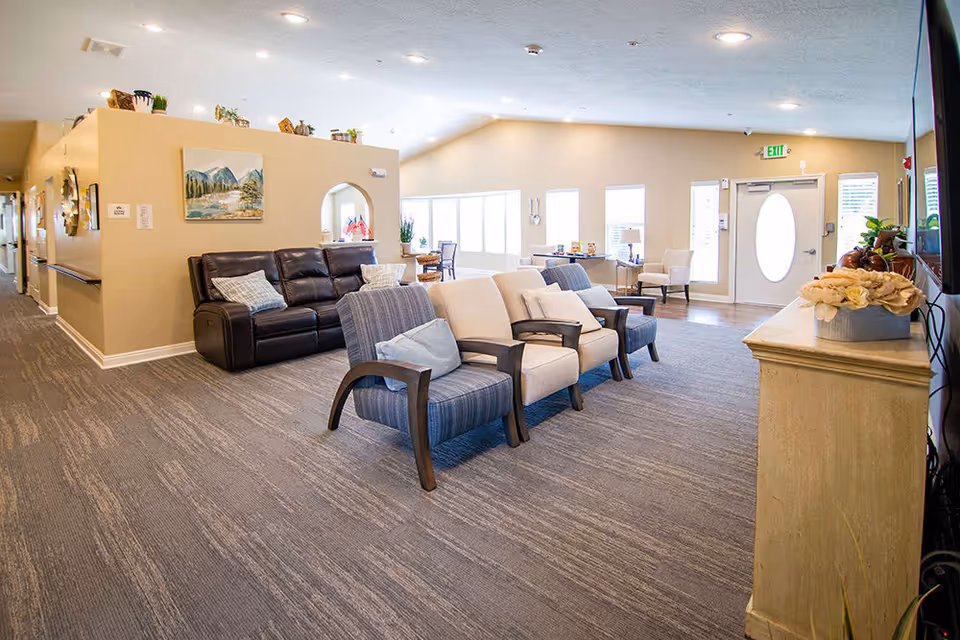 A bright and spacious common area in a senior living facility featuring a row of cushioned chairs with armrests, a dark leather sofa with pillows, a light-colored cabinet with a flower arrangement, and large windows letting in natural light. The walls are painted beige and decorated with paintings and small plants on top of a half wall.