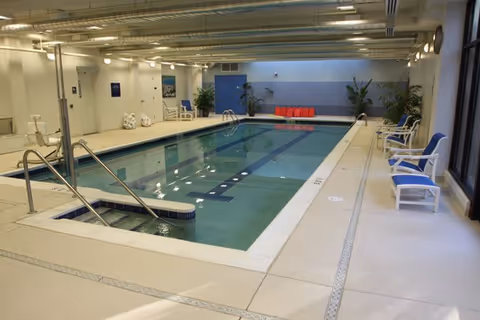 Indoor swimming pool with lounge chairs, potted plants, and handrails inside a retirement community facility.