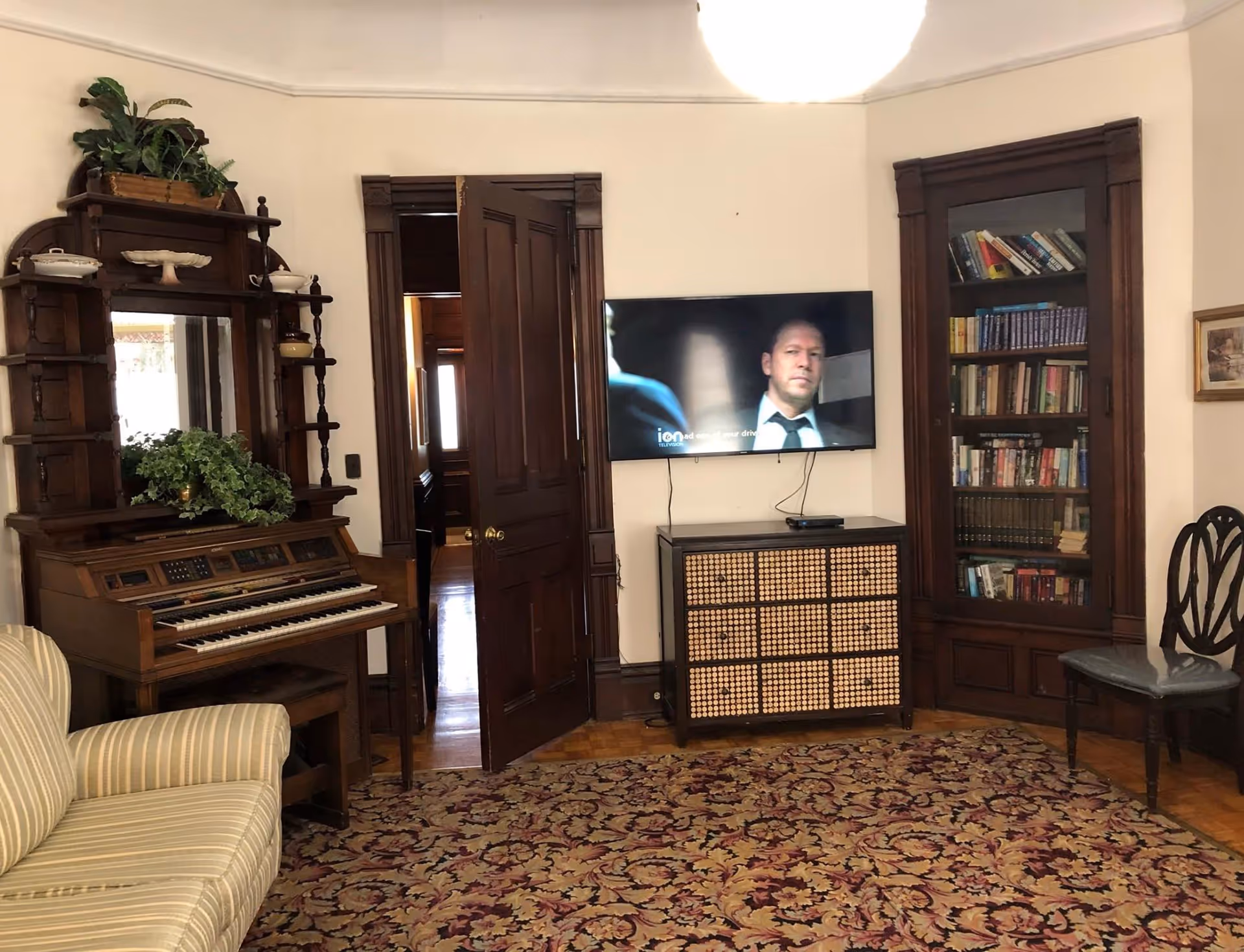 A cozy living room with a striped sofa, an antique wooden organ with plants on top, a flat-screen TV mounted on the wall showing a man in a suit, a wooden cabinet with a woven front, a built-in wooden bookshelf filled with books, a wooden chair with a cushioned seat, and a patterned area rug covering the floor.