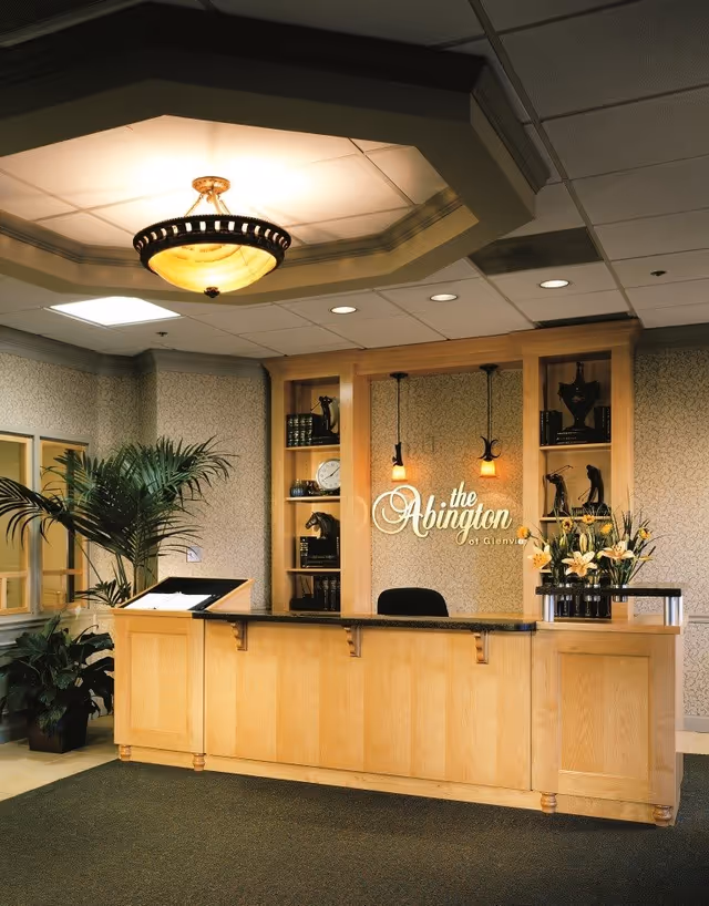 Reception area with a wooden front desk, decorative shelves with books and sculptures, a large ceiling light fixture, potted plants, and a sign on the wall that reads 'the Abington of Glenvie'.