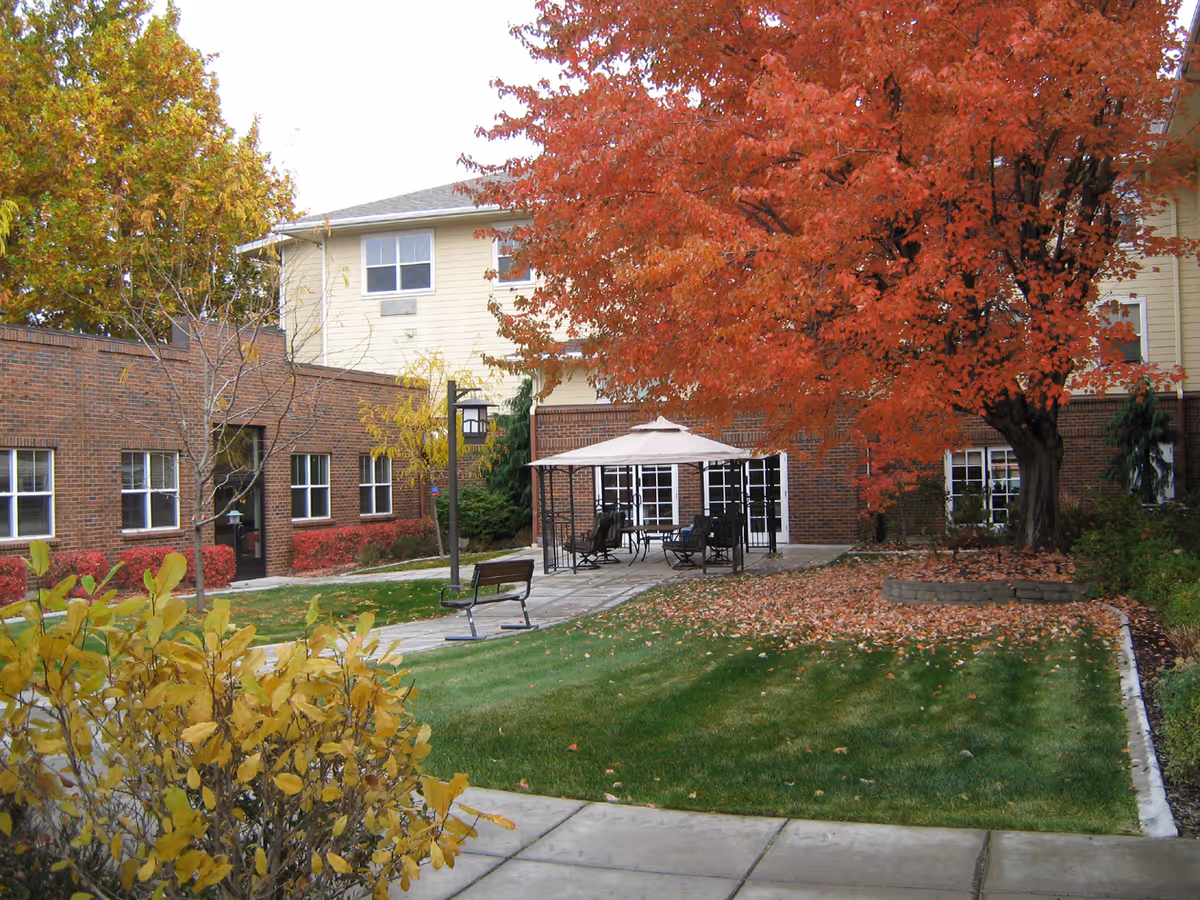 Outdoor courtyard area at a senior living facility with a large tree with red autumn leaves, a paved walkway, a bench, and a covered seating area with chairs and a table. The building surrounding the courtyard has brick and light-colored siding with multiple windows.