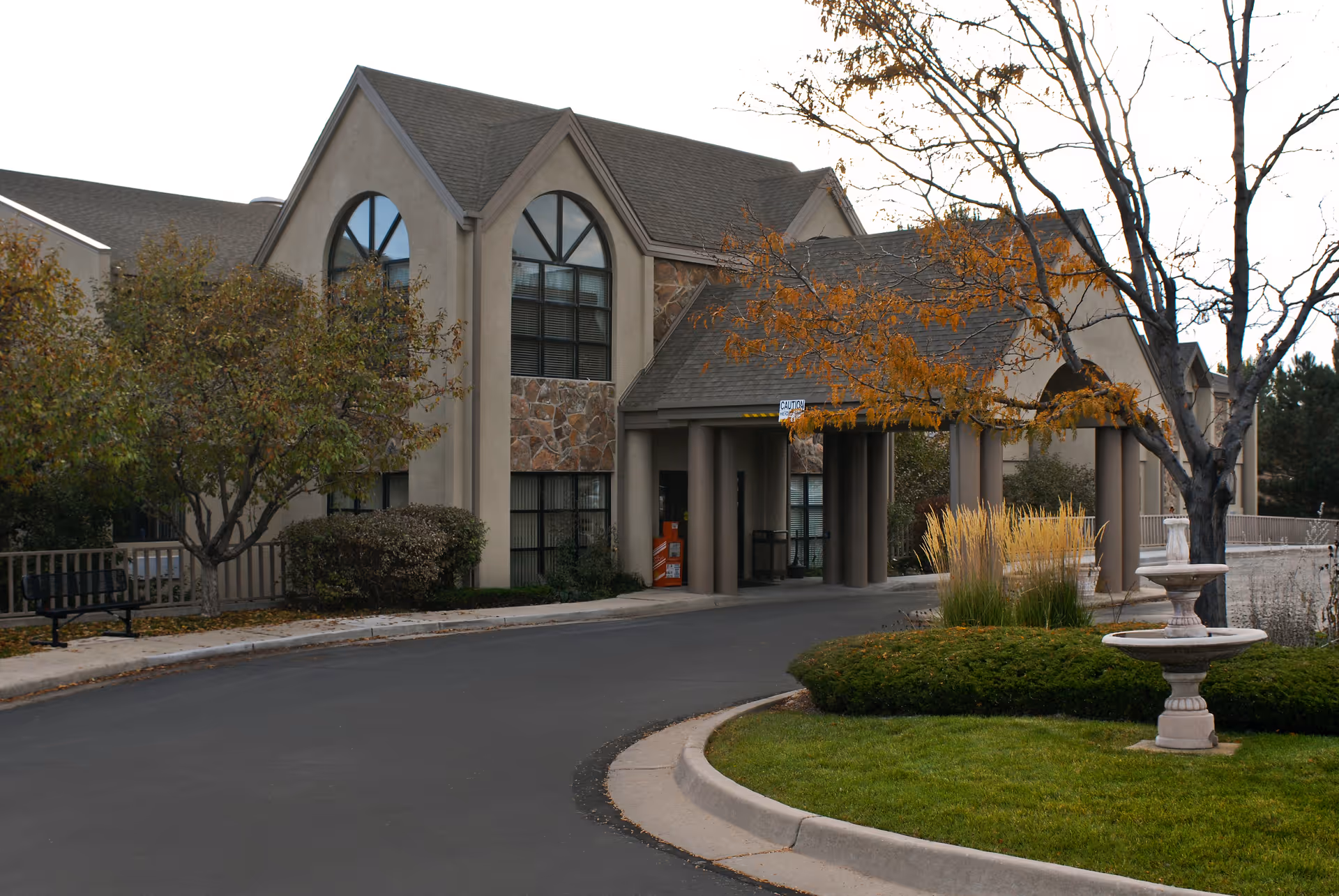 Exterior view of a senior living facility building with a covered entrance, arched windows, and a landscaped area with trees and a fountain in front.