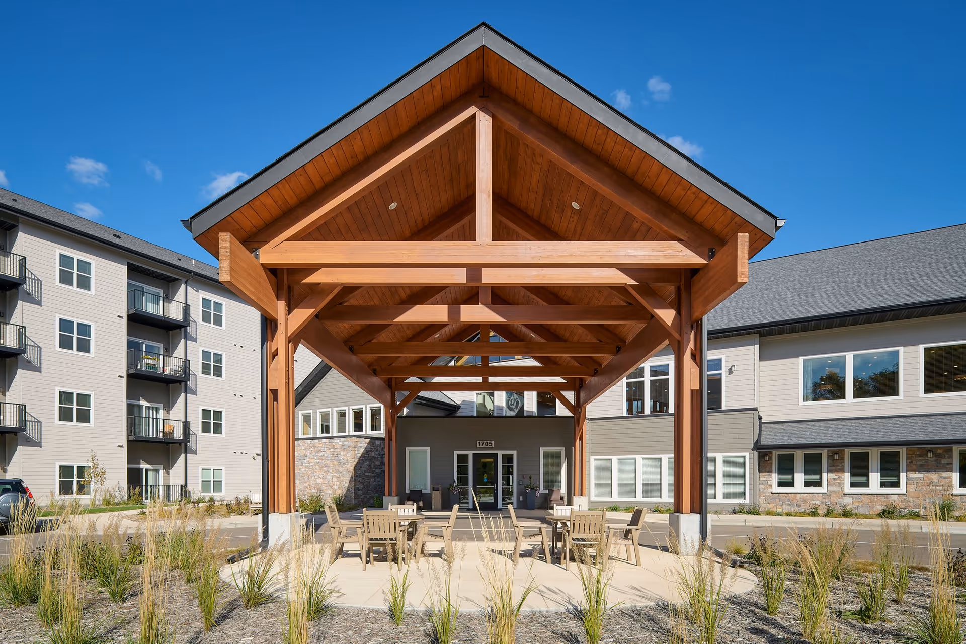 Outdoor covered seating area with wooden beams and a peaked roof in front of a multi-story senior living facility building under a clear blue sky. Several tables and chairs are arranged under the structure, surrounded by landscaped plants and grasses.