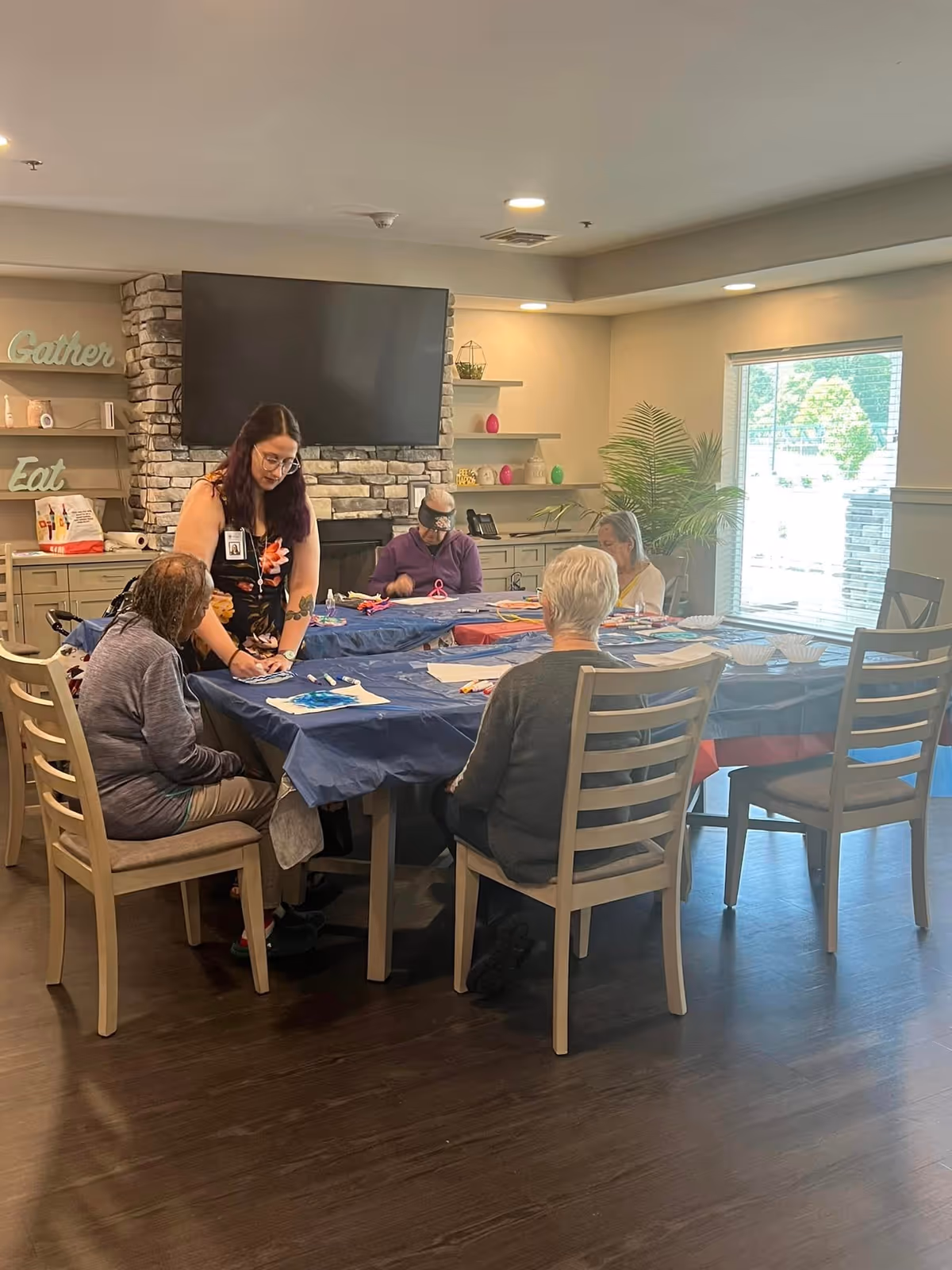 A group of elderly people sitting around a table covered with a blue tablecloth, engaging in an activity with a caregiver standing and assisting them. The room has a stone fireplace with a large TV mounted above it, shelves with decorative items, and a large window letting in natural light.