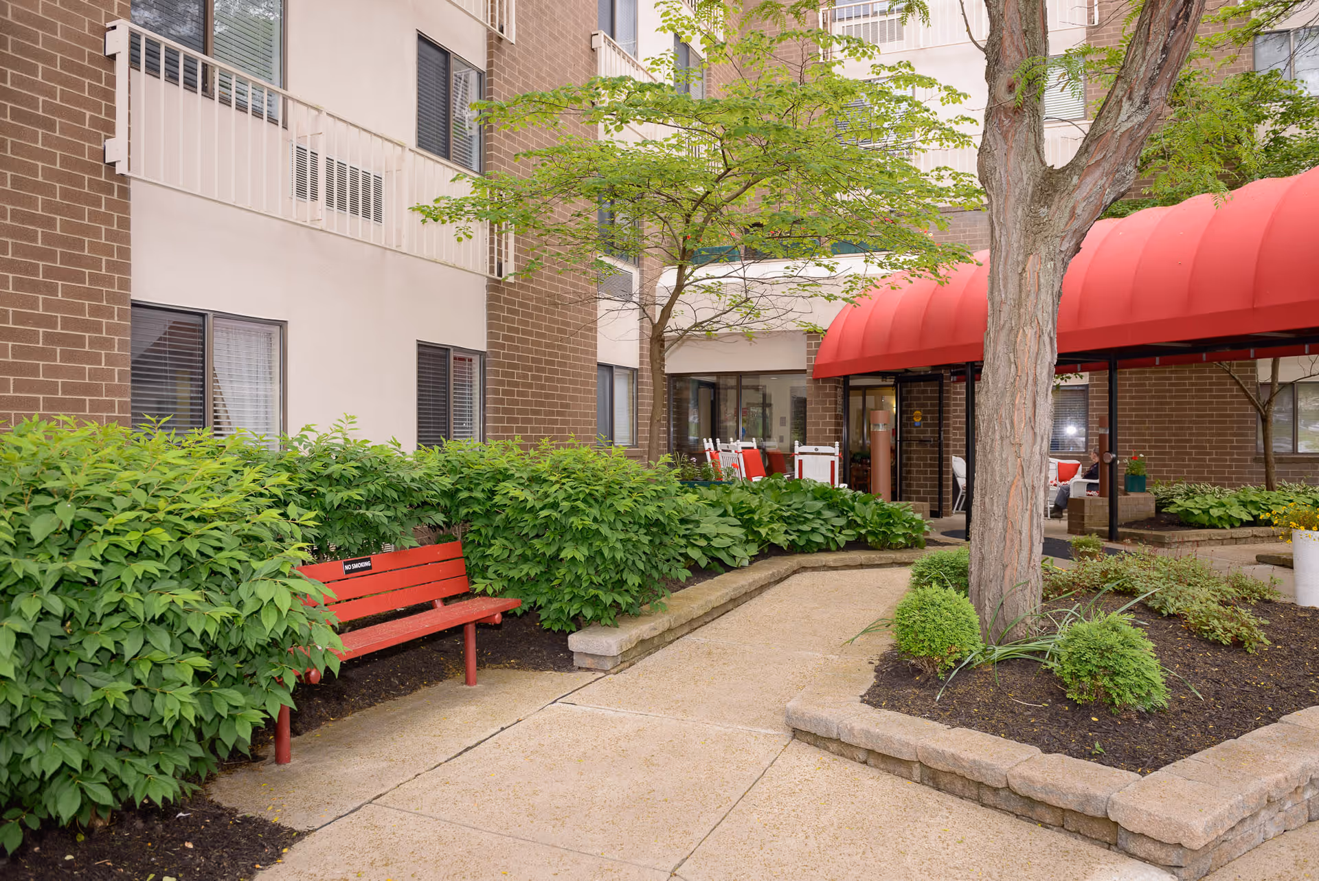 Outdoor courtyard area of a senior living facility with a red bench, green bushes, a tree, and a red awning over a seating area near the building entrance.