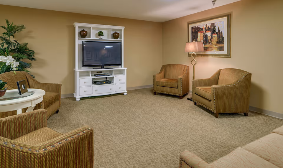 A cozy living room with beige walls and carpet, featuring a white entertainment center with a flat-screen TV, three brown upholstered armchairs, a round white side table with a framed photo and a potted orchid, a floor lamp, a framed painting on the wall, and a large green plant in the corner.