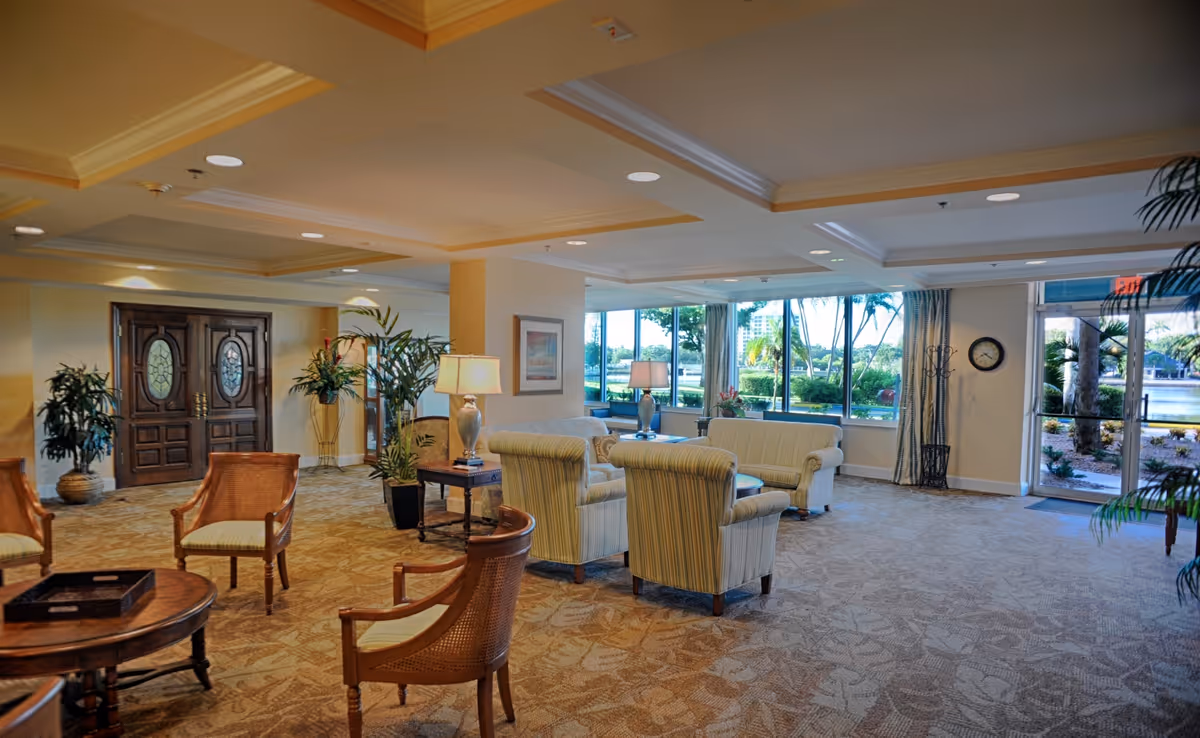 A spacious and well-lit living room area with multiple upholstered armchairs and sofas arranged around wooden tables. The room features large windows with views of greenery and water outside, beige walls, patterned carpet, ceiling lights, and decorative plants. There is a wooden double door on one side and an exit door on the other side.