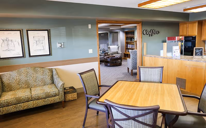 A cozy senior living common area with a wooden table and four chairs in the foreground. To the left, there is a patterned sofa against a wall with two framed sketches. In the background, there is an open doorway leading to another room with more seating and bookshelves. On the right side, there is a kitchen counter with a sign that says 'Coffee' on the wall above it.