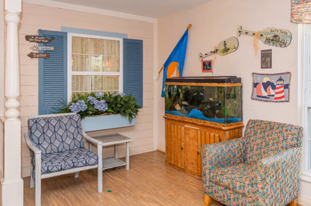 A cozy interior corner featuring two upholstered chairs with different patterns, a small wooden side table, a large fish tank on a wooden cabinet, and decorative wall hangings including a blue flag, paddle signs, and a nautical-themed tapestry. There is a window with blue shutters and a flower box filled with purple flowers and greenery.