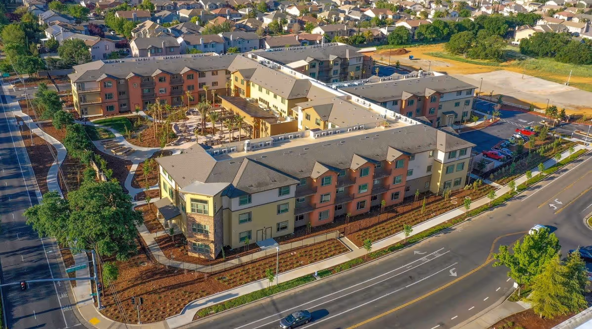 Aerial view of Bruceville Point, a large multi-story residential building complex surrounded by landscaped gardens, walkways, parking lots, and roads. The complex is situated in a suburban neighborhood with many houses in the background.