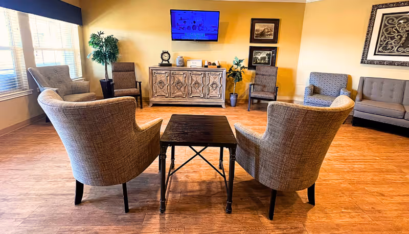 Communal lounge with upholstered armchairs around a small table facing a wall-mounted TV and decorative cabinet.
