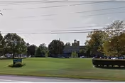 A wide grassy lawn with trees and hedges in front of a low institutional building and parked cars beneath overhead power lines.
