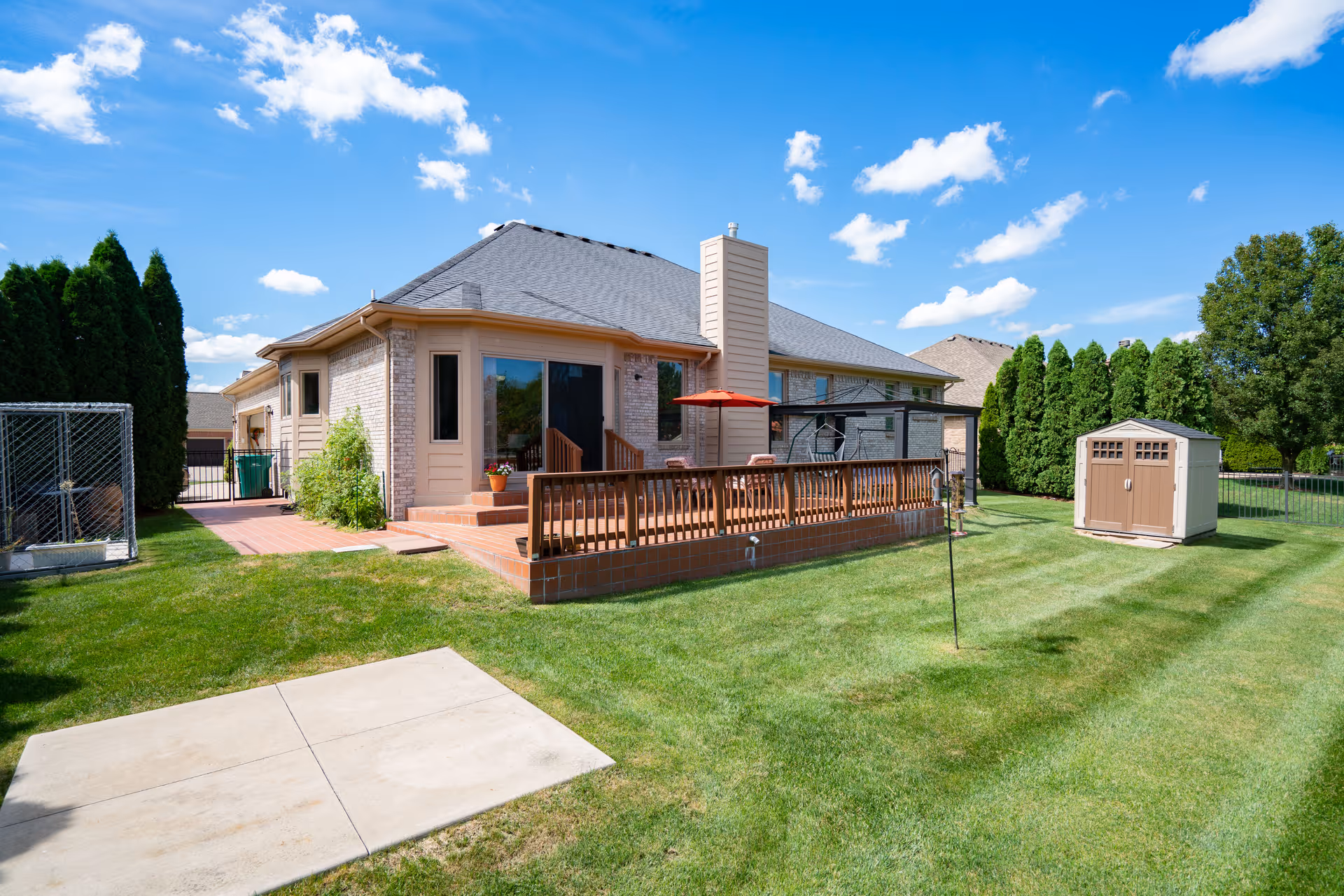A backyard view of a single-story brick house with a wooden deck featuring outdoor furniture and a red umbrella. The yard is well-maintained with green grass, a small storage shed, tall evergreen trees along the fence, and a clear blue sky with scattered clouds.