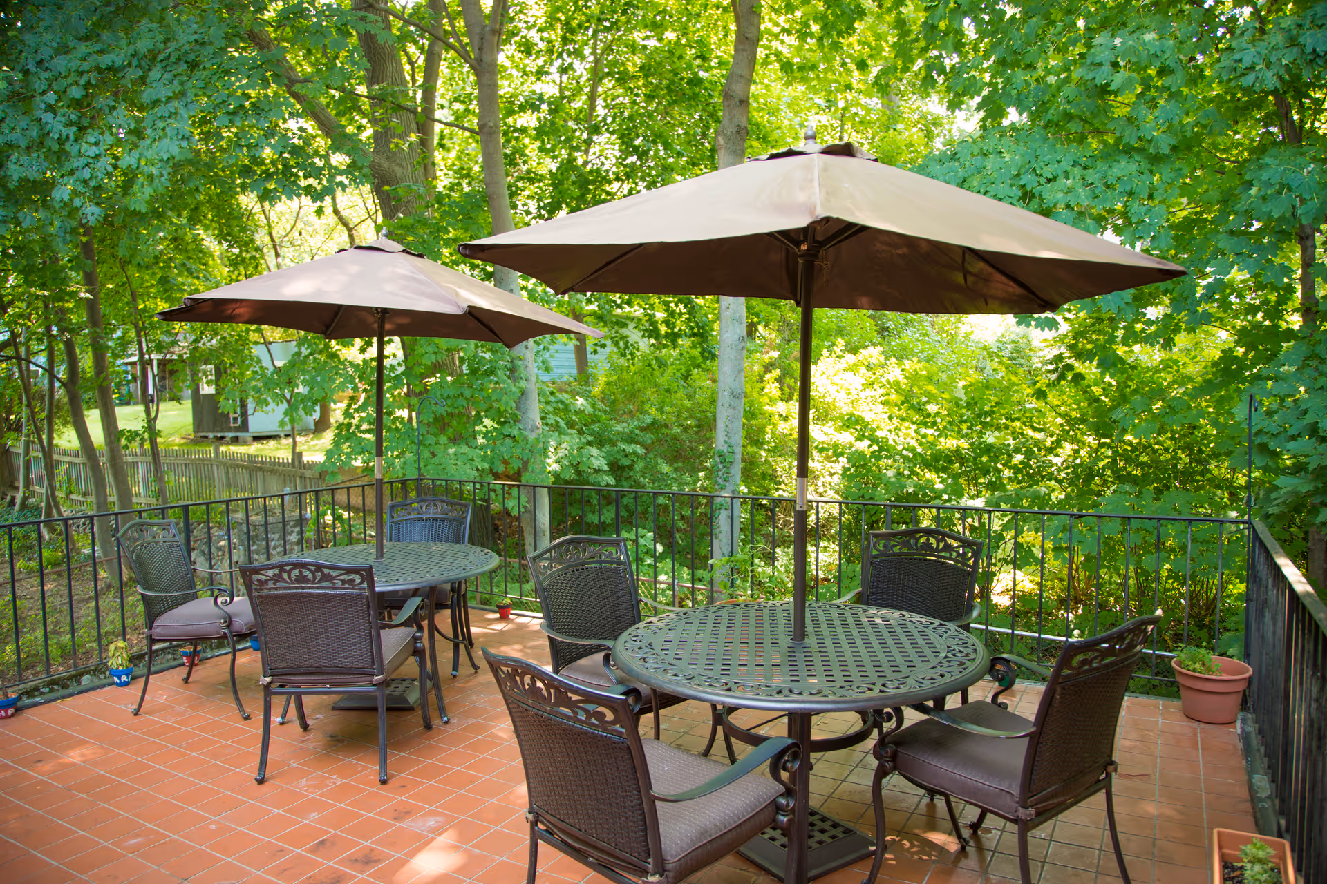 Outdoor patio with metal tables, chairs, and large umbrellas surrounded by green trees.