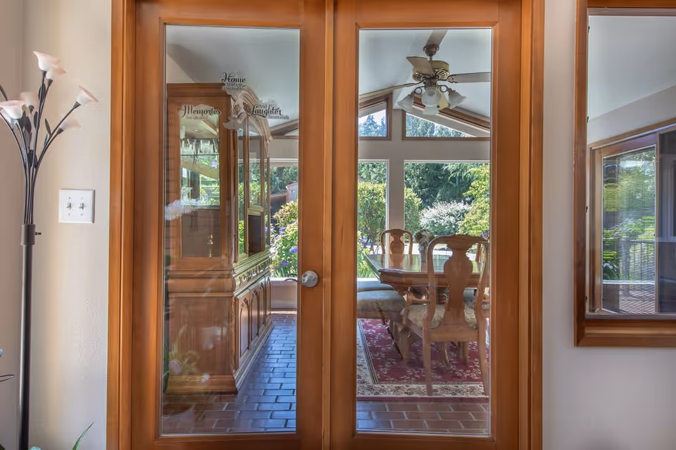 View through glass double doors into a dining room with a wooden dining table and chairs on a red patterned rug. The room has large windows with a view of green trees and bushes outside. A wooden cabinet is visible on the left side inside the dining room, and a ceiling fan with lights hangs from the ceiling. A tall floor lamp is positioned to the left of the doors.