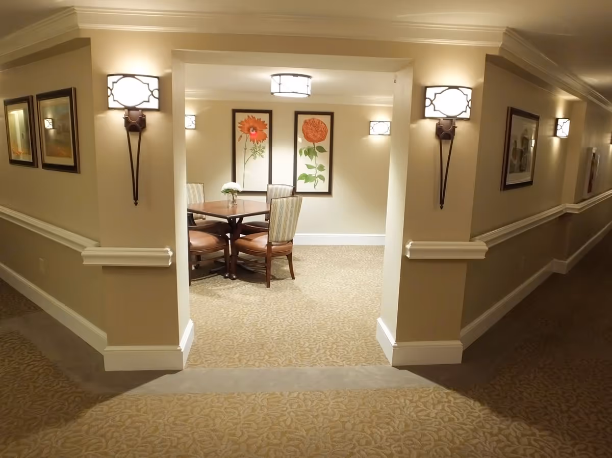 A well-lit interior hallway leading to a small dining area with a round wooden table and four chairs. The walls are beige with white trim, decorated with framed floral artwork and wall sconces providing soft lighting. The carpet has a subtle patterned design.