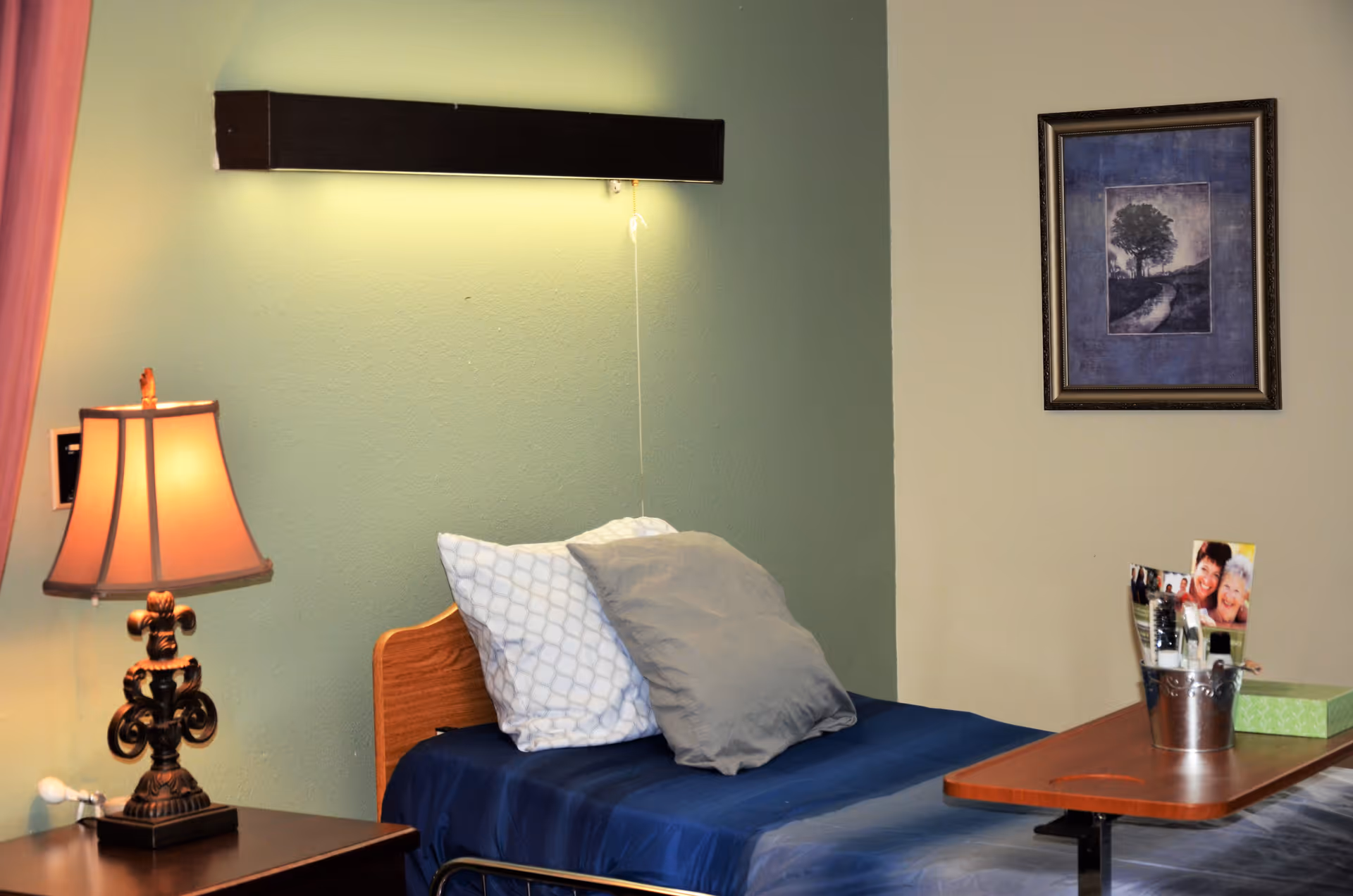 A single bed with blue bedding and two pillows in a room with green and beige walls. There is a wooden nightstand with an ornate lamp on it next to the bed. Above the bed is a wall-mounted light fixture with a pull cord. A framed picture hangs on the beige wall, and a small table with personal items and photos is positioned next to the bed.