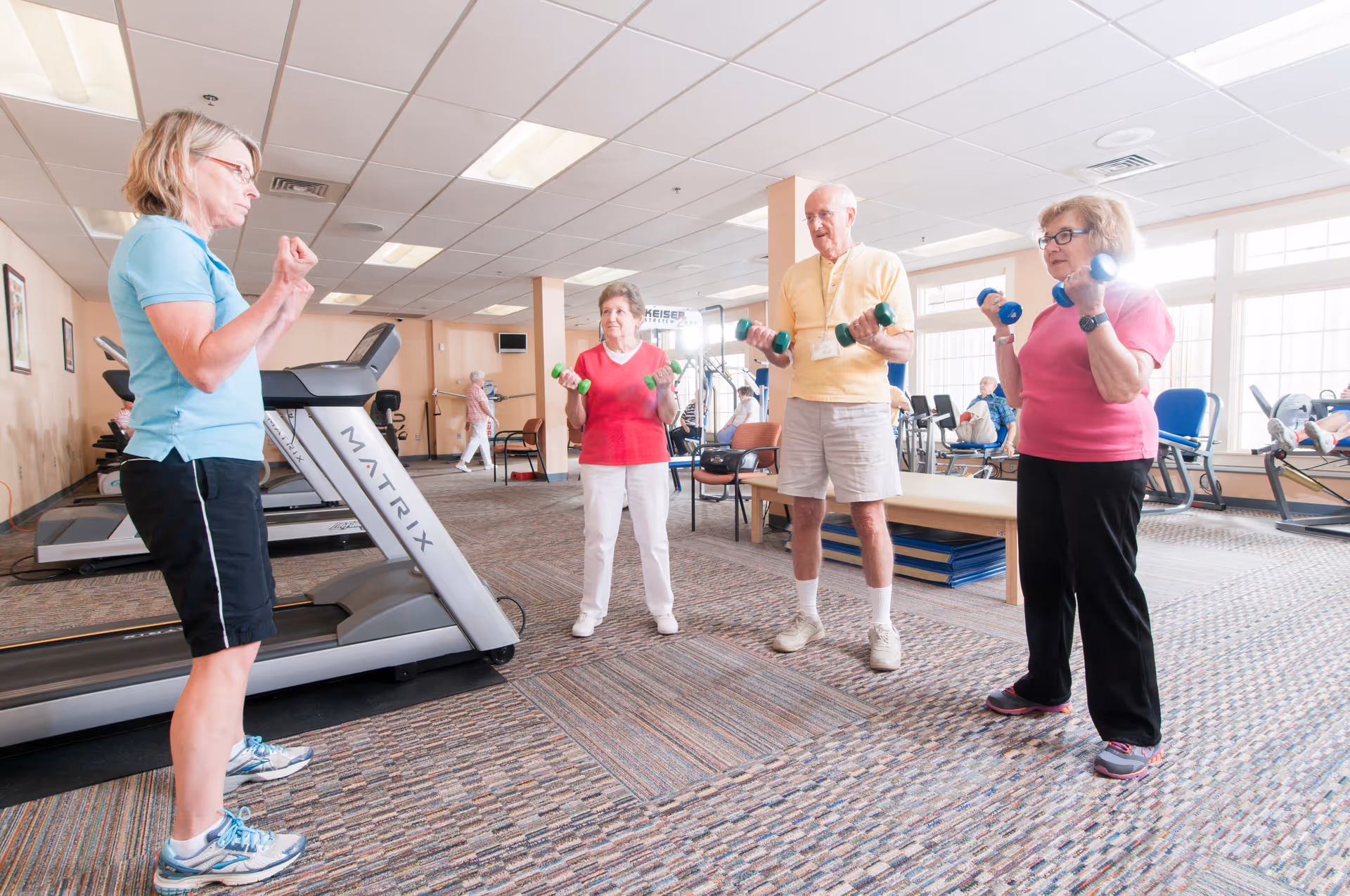 A group of three elderly individuals lifting small dumbbells in a fitness room, guided by a female instructor. The room has treadmills, exercise equipment, and large windows letting in natural light.