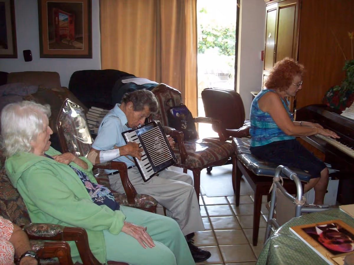 A group of elderly people in a living room setting. One elderly man is playing an accordion while a woman is playing a piano. Other elderly individuals are seated nearby, watching and listening. The room has chairs, a table with a book or magazine, and a window with curtains letting in natural light.