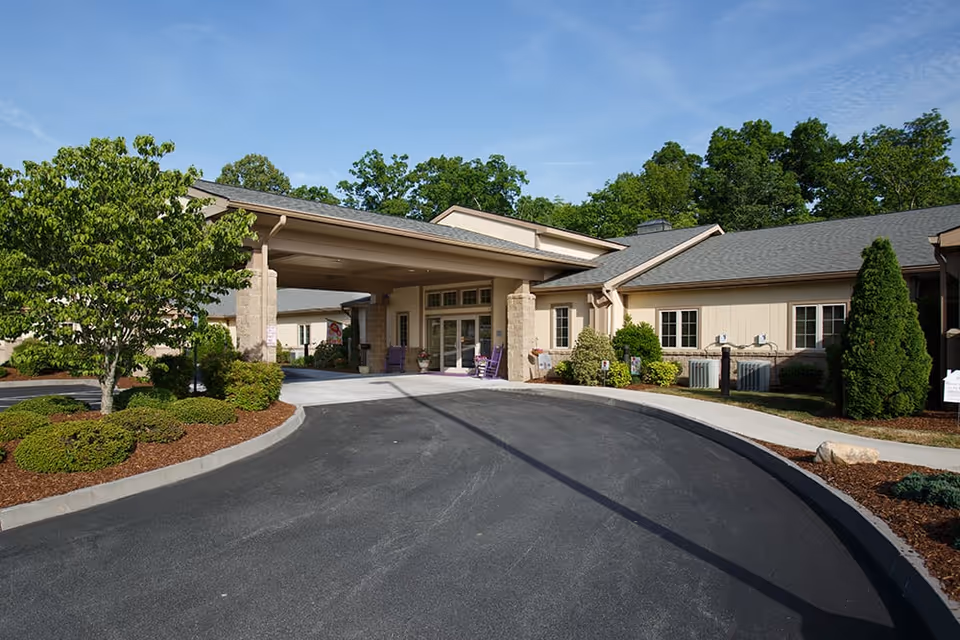 Exterior view of Preston Place II facility entrance with a covered drop-off area, surrounded by landscaped bushes, trees, and a paved driveway under a clear blue sky.