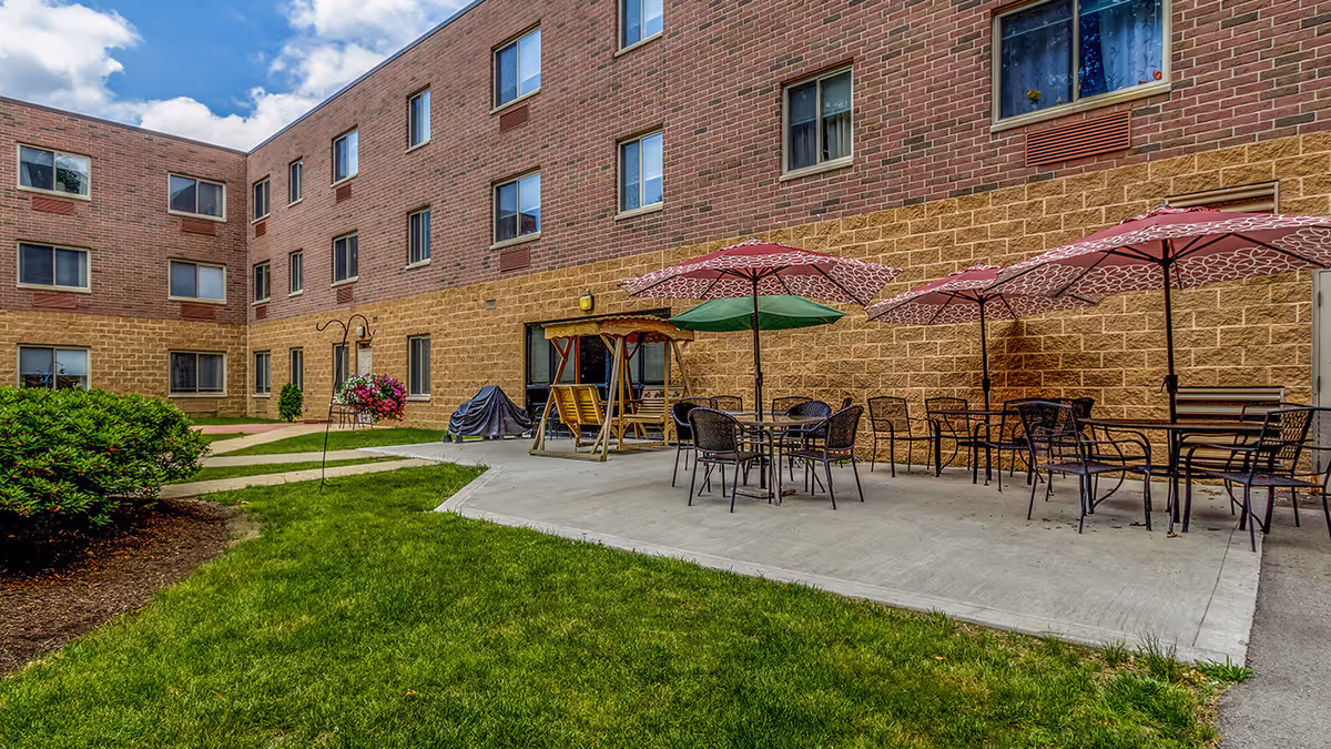 Outdoor patio area at Whispering Oaks - A Provincial Senior Living Community featuring several tables with chairs and umbrellas, a wooden swing, green grass, bushes, and a multi-story brick building in the background under a partly cloudy sky.