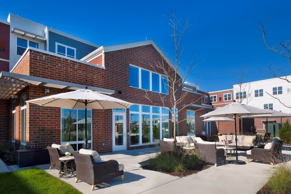 Outdoor patio area of a senior living facility with brick exterior walls, large windows, and multiple seating arrangements including cushioned chairs and sofas under large white umbrellas. The sky is clear and blue, and there are small trees and landscaped greenery around the patio.