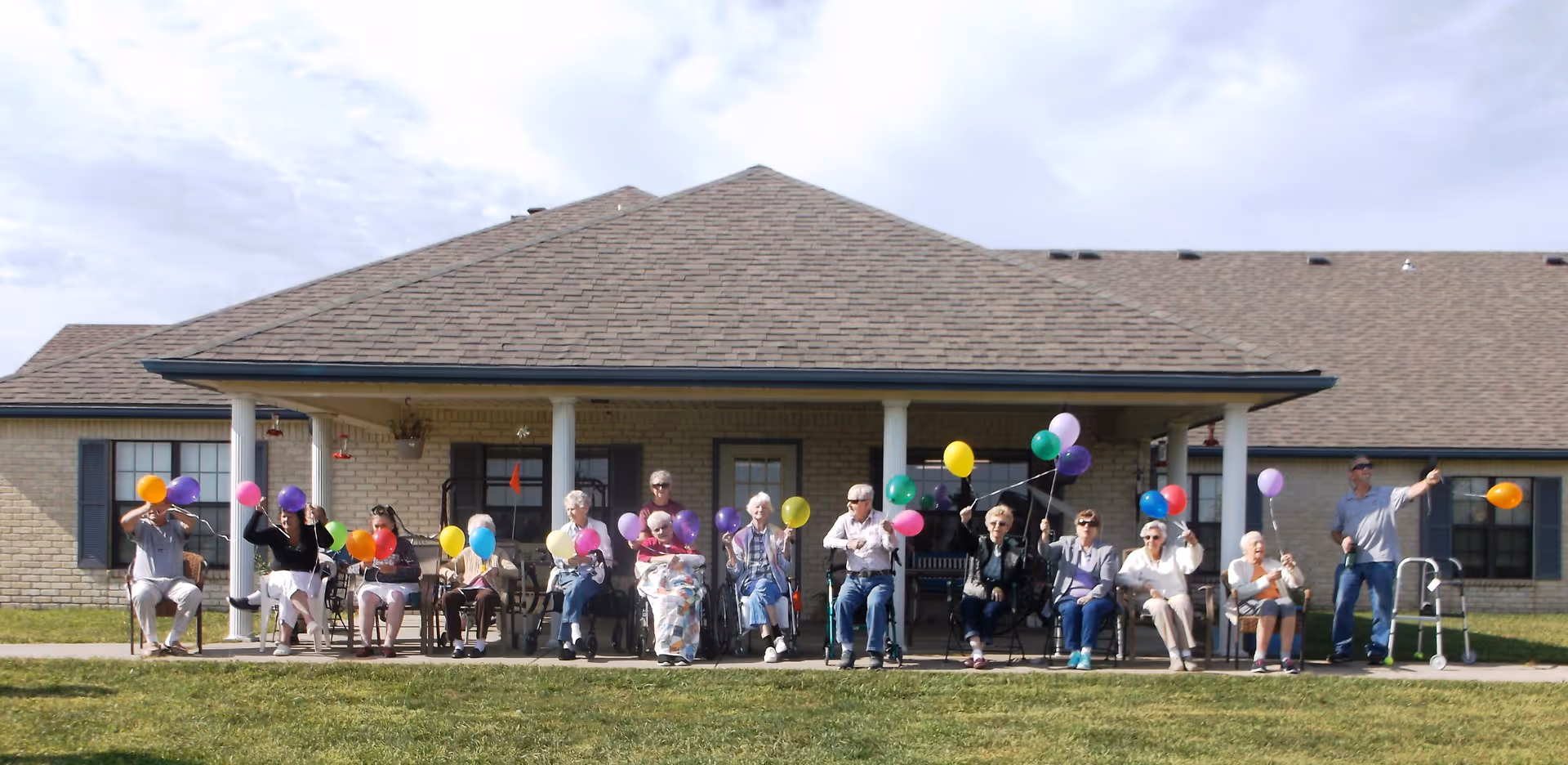 A group of elderly people sitting on chairs and wheelchairs on a porch in front of a single-story brick building, each holding colorful balloons. The sky is partly cloudy and the grass is green in front of them.
