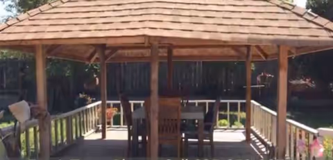 A wooden gazebo with a shingled roof situated outdoors, containing a table and several chairs inside. The gazebo is surrounded by a wooden railing and greenery in the background.