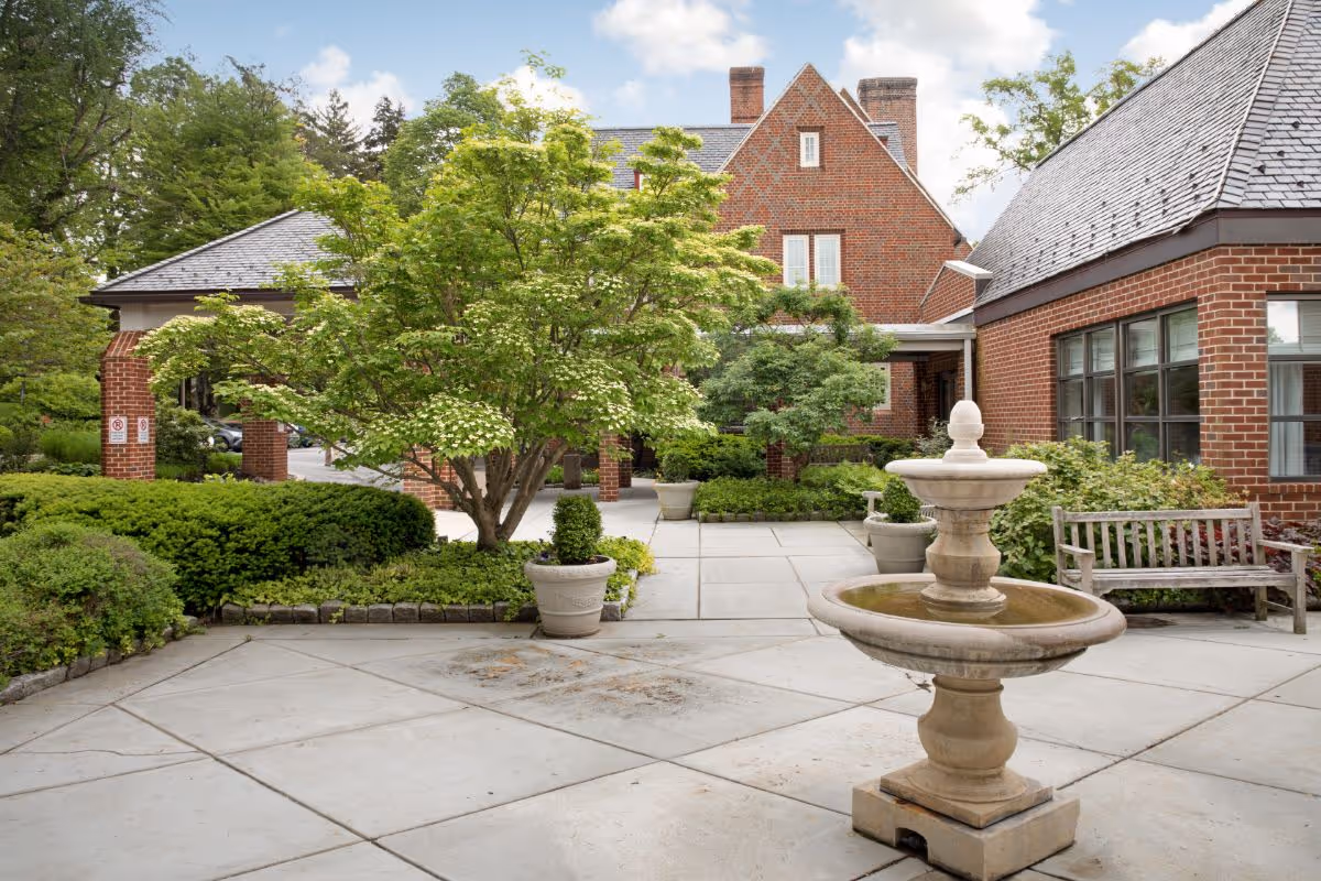 Outdoor courtyard area at The Quadrangle featuring a stone fountain in the foreground, surrounded by paved walkways, green shrubs, potted plants, a wooden bench, and brick buildings with large windows under a partly cloudy sky.