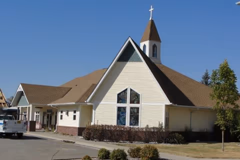 Exterior view of a senior living community building with a steeply pitched roof and a small steeple topped with a cross, beige siding, and large triangular windows. There is a driveway with a white truck parked on the left and some landscaping with bushes and trees around the building under a clear blue sky.