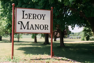 A large white sign with red posts displaying the text 'Leroy Manor' in black letters, set in a grassy area with trees and a clear sky in the background.