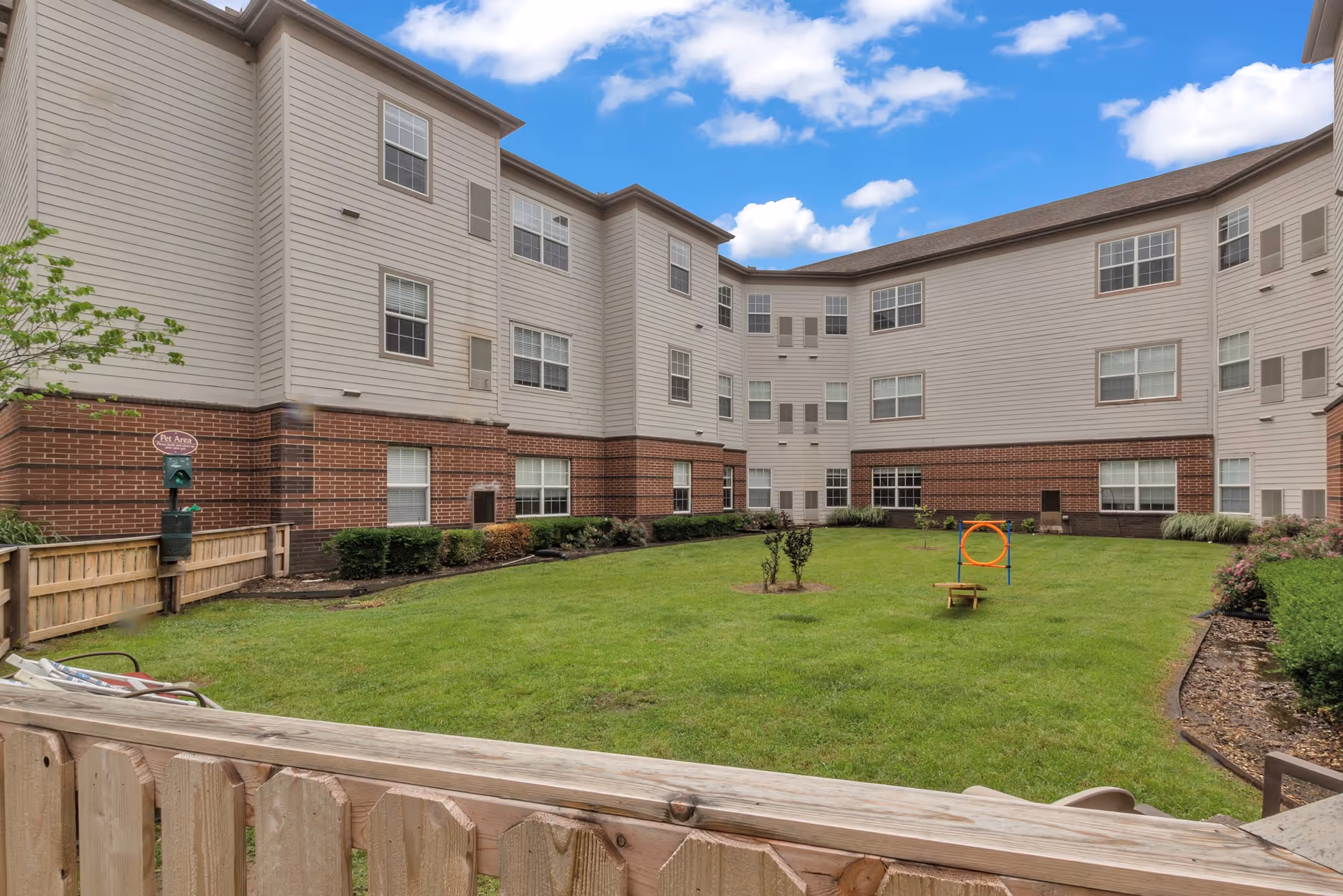 A grassy courtyard area surrounded by a three-story residential building with beige siding and red brick lower walls. The courtyard has a small fenced pet area with a pet waste station and a dog agility hoop. The sky is blue with scattered clouds.