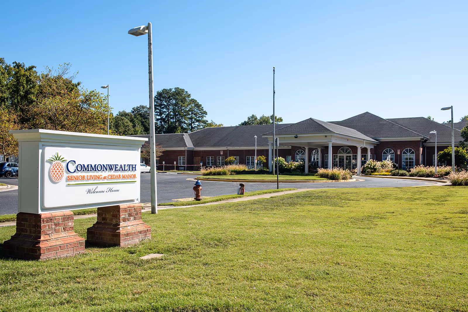 Exterior view of Commonwealth Senior Living at Cedar Manor, showing a brick building with a covered entrance and arched windows, a large green lawn, a paved driveway, and a sign with the facility's name and logo in the foreground.