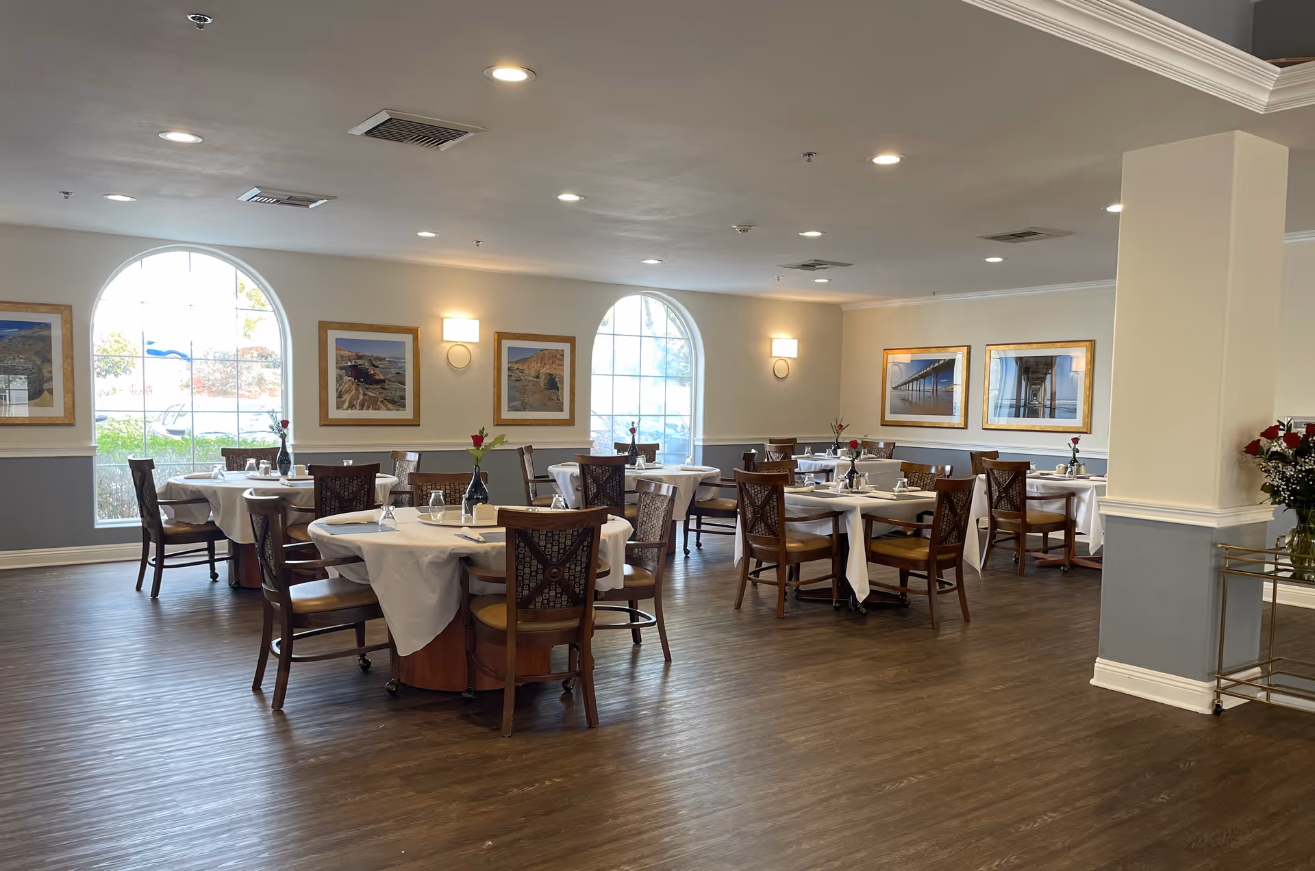 Bright, spacious dining room with round tables set with white tablecloths and wooden chairs, arched windows and framed artwork on the walls.
