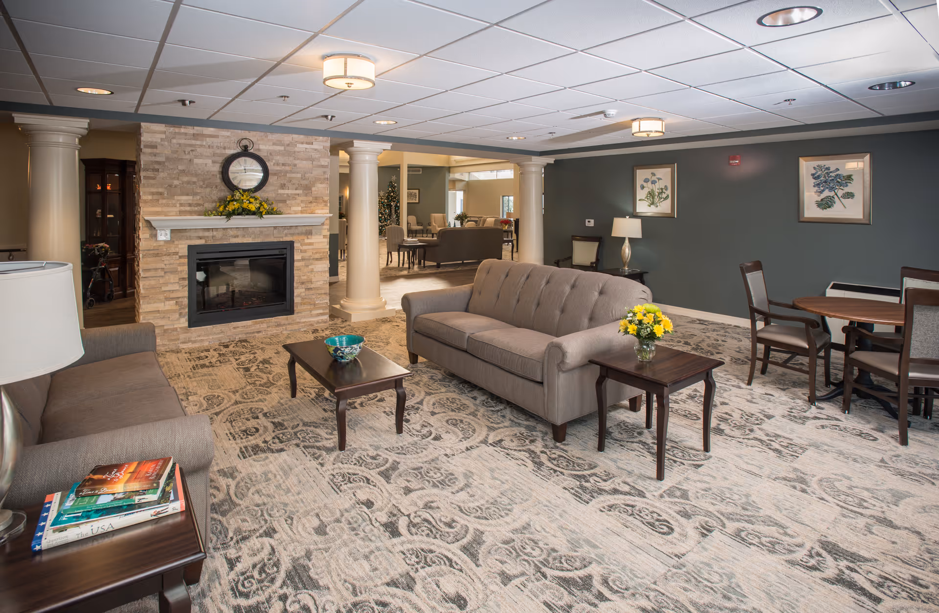 A cozy senior living common lounge with sofas, coffee tables, a fireplace flanked by columns, and a dining table in the background.