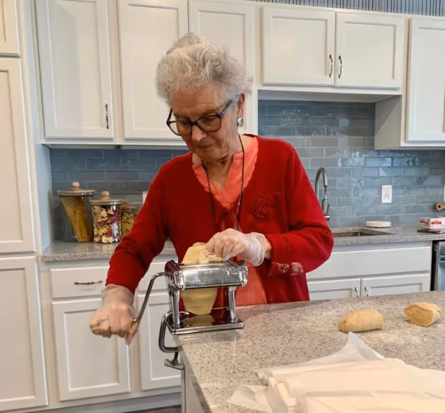 An elderly woman wearing glasses and a red sweater is using a pasta machine to roll out dough in a modern kitchen with white cabinets and a gray tiled backsplash.