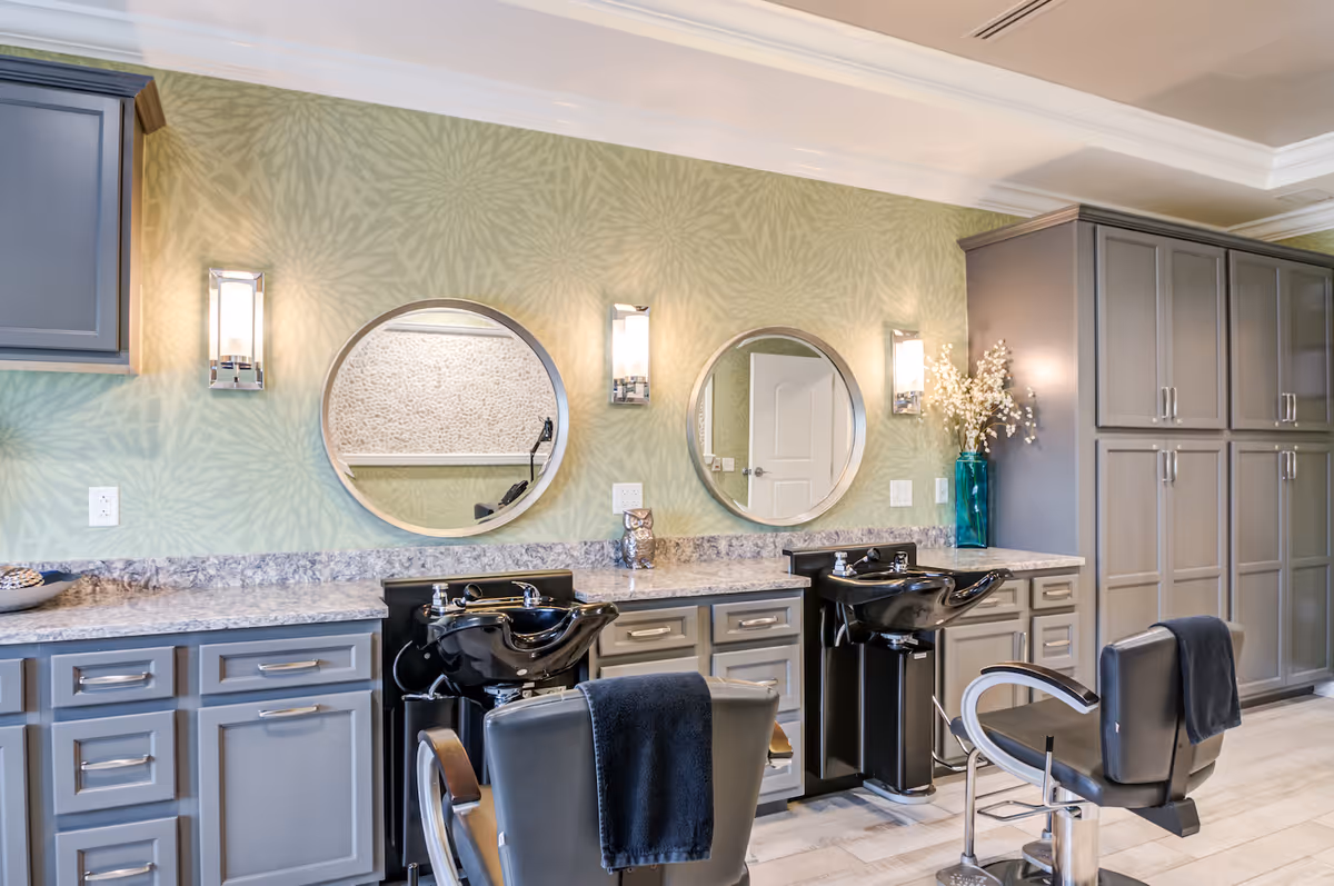 Interior view of a salon area with two black salon chairs facing two black wash basins. The wall behind has a green patterned wallpaper with two round mirrors and two wall-mounted lights. There are gray cabinets and drawers below the countertop and a tall cabinet on the right side. A blue vase with white flowers is placed on the countertop.