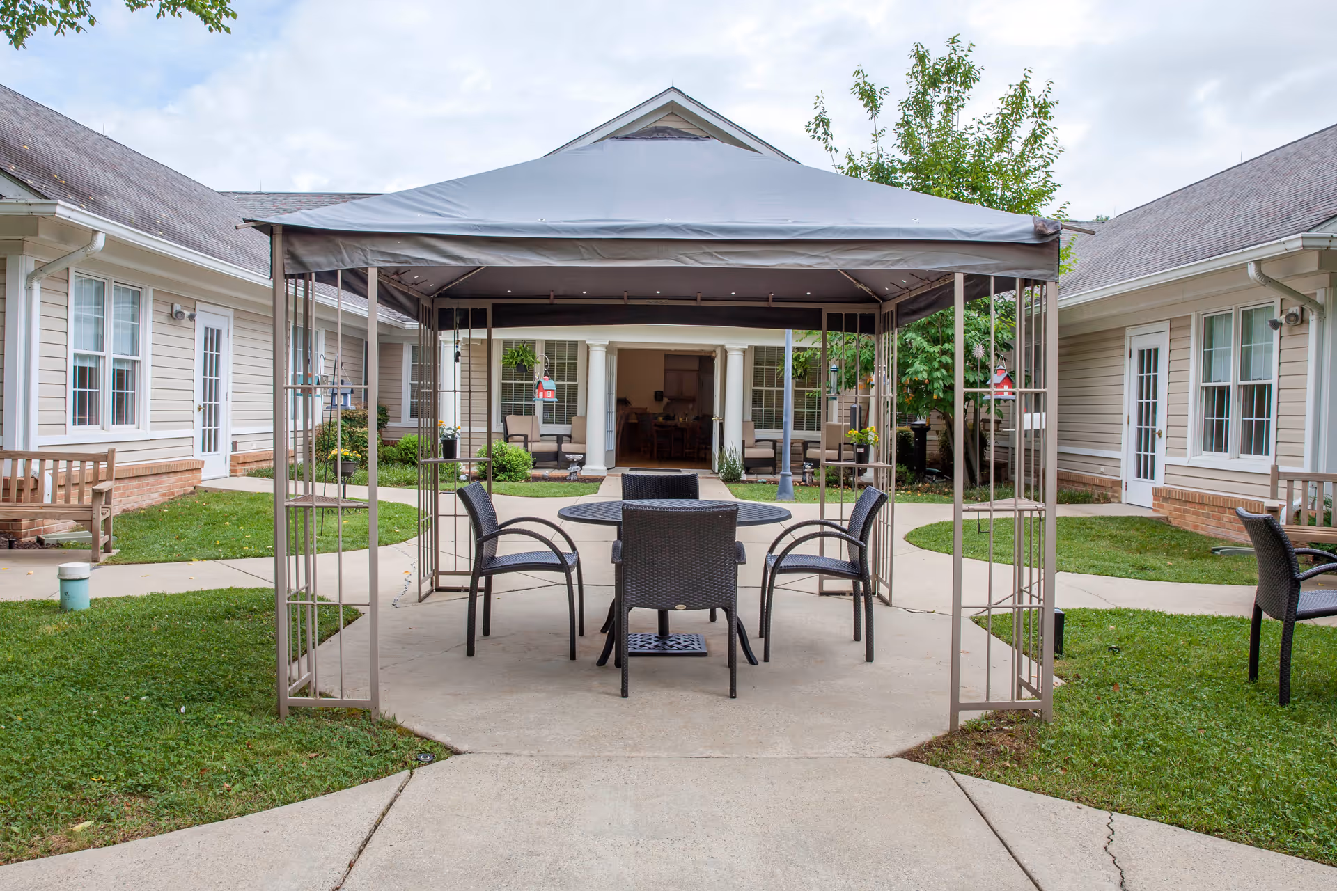 Outdoor seating area with a metal gazebo covering a round table and four chairs, surrounded by a courtyard with grass, pathways, benches, and buildings with beige siding and white trim.