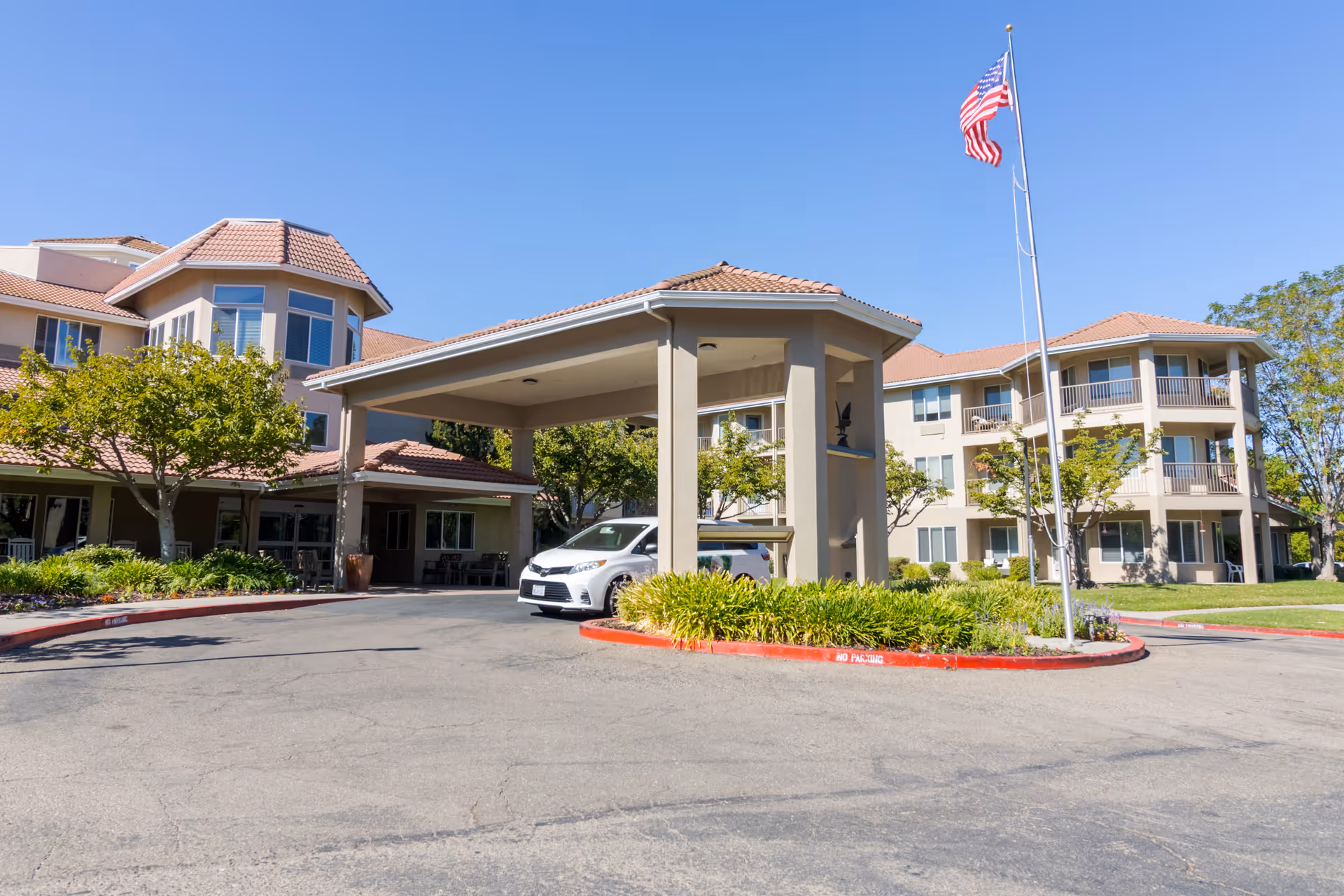 Exterior view of Quail Lodge Retirement Community building with a covered entrance, a white car parked under the canopy, landscaped greenery, and an American flag on a flagpole against a clear blue sky.