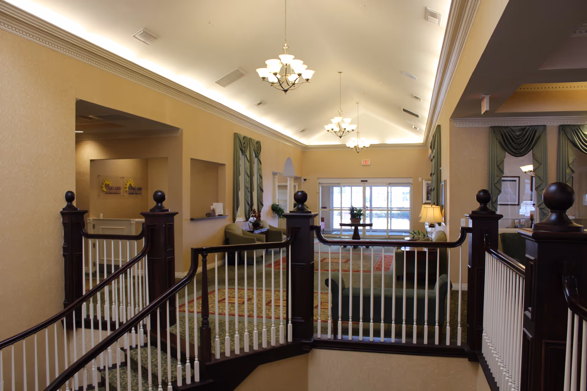 Interior view of a senior living facility lobby area with a staircase railing in the foreground, chandeliers hanging from a high ceiling, seating areas with sofas and chairs, decorative curtains on the windows, and a glass entrance door at the far end.